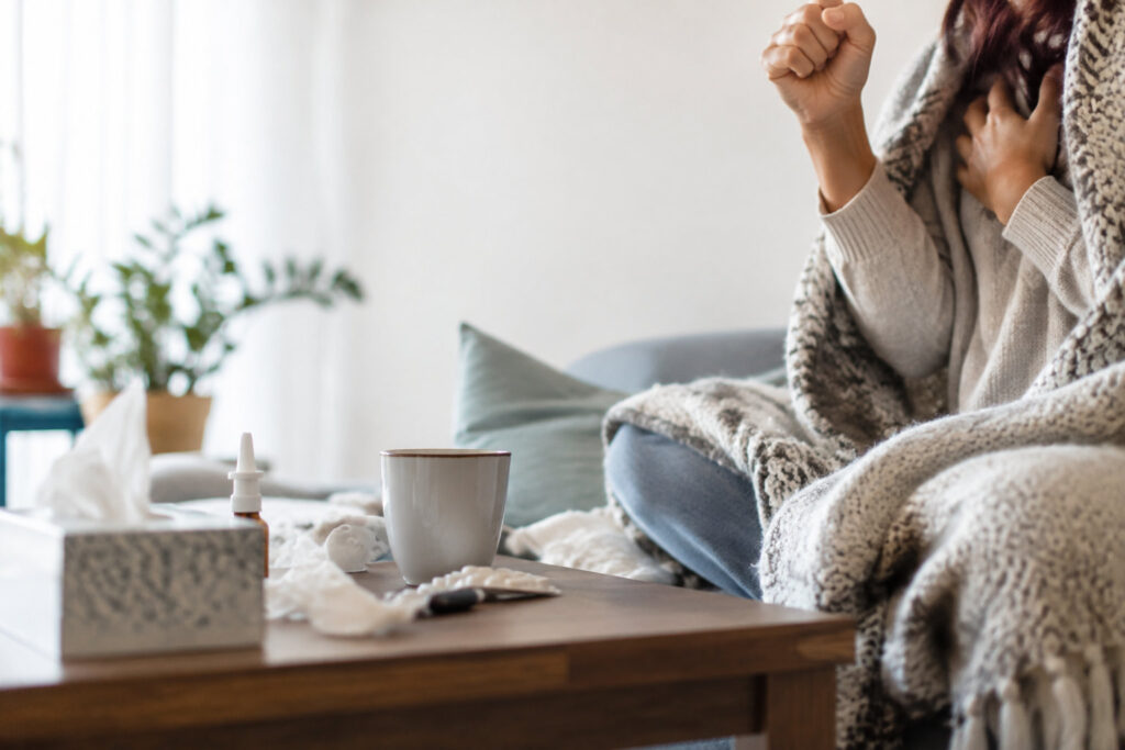 Une personne malade tousse sous une couverture, avec des médicaments et une boisson chaude sur une table devant elle.